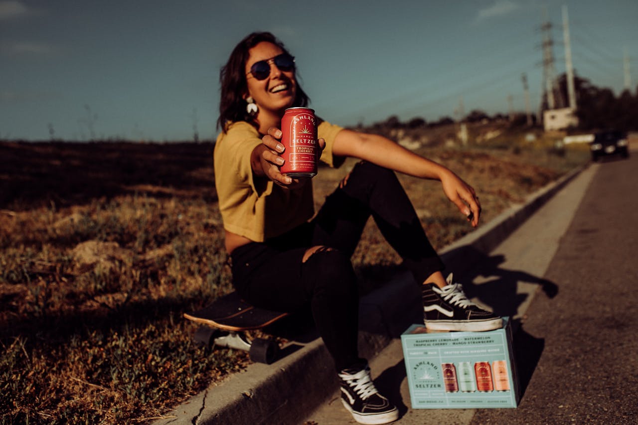 services-01 Woman enjoying a canned beverage outdoors, sitting on a skateboard with a big smile.